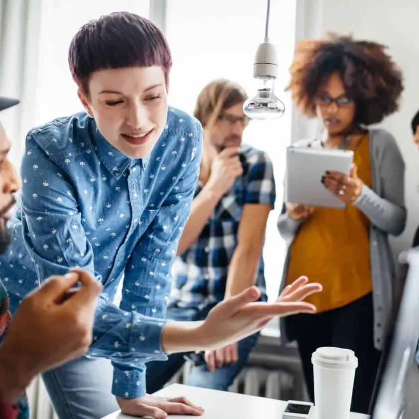 Two young start-up business people discussing at computer monitor their latest project. Their colleagues are visible while standing in the background and holding a digital tablet.